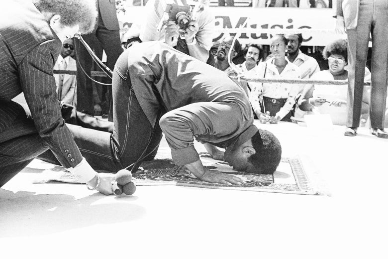 A kneeling Ali rests his head on a prayer mat in a boxing ring while surrounded by press.